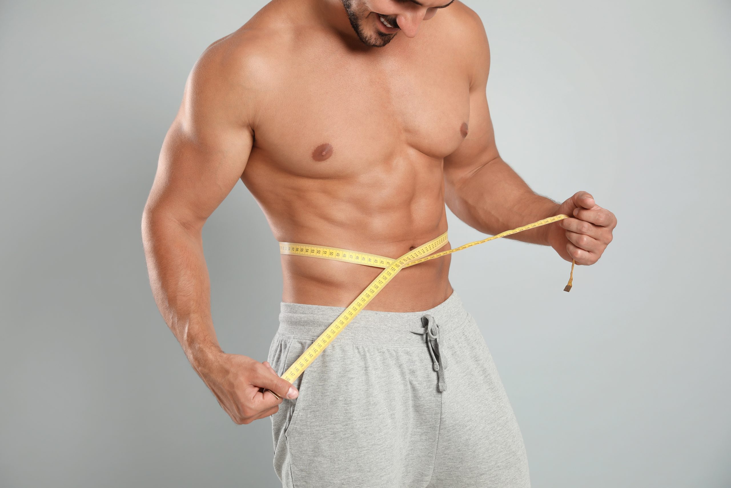 Young man with slim body using measuring tape on grey background, closeup view